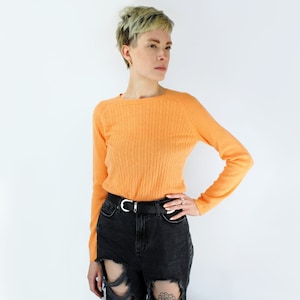 May include: A peach-colored, long-sleeved sweater with a cable knit pattern. The sweater is worn with black jeans and a black belt.