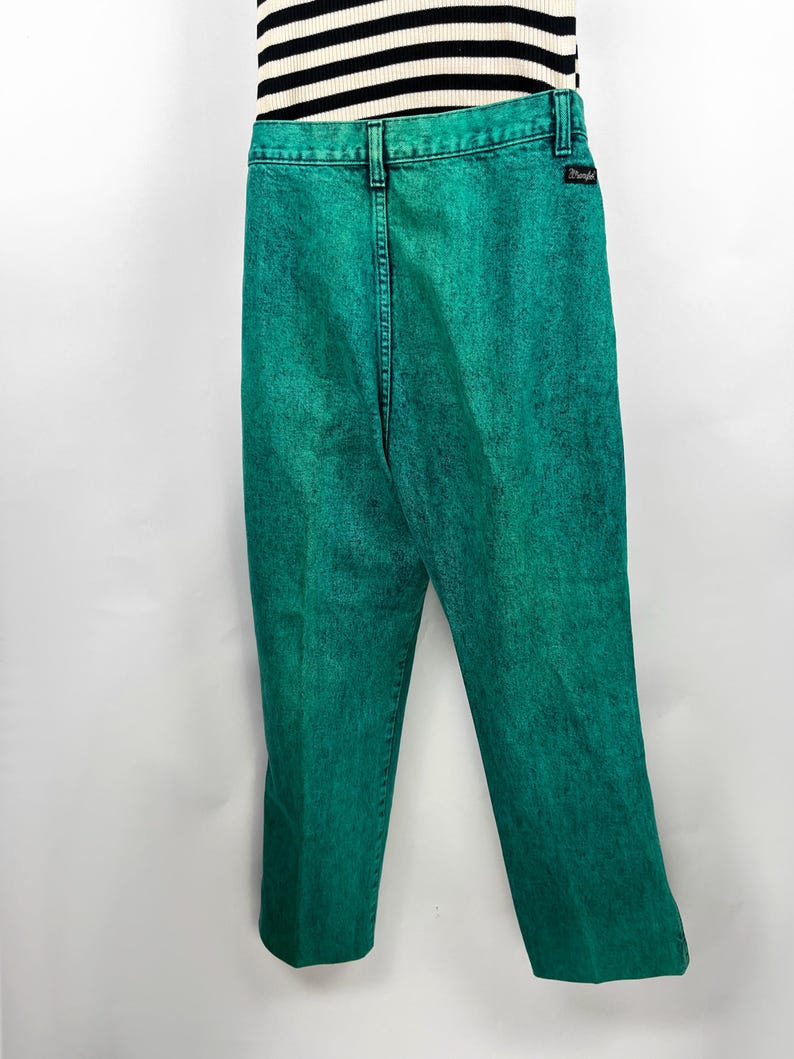 May include: Bright green acid wash jeans with a black Wrangler label on the back pocket. The jeans are paired with a black and white striped top. The pants have a classic five-pocket design and a straight leg.