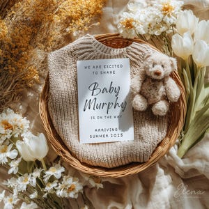 May include: A beige baby sweater rests in a woven basket, alongside a small teddy bear and a card announcing "Baby Murphy is on the way, arriving Summer 2025." The scene is decorated with white flowers and dried foliage, creating a soft, neutral aesthetic.