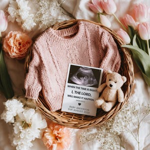 May include: A pink cable-knit baby sweater, a sonogram image, and a small teddy bear are arranged in a woven basket, surrounded by flowers. The sonogram card reads "WHEN THE TIME IS RIGHT, I, THE LORD, WILL MAKE IT HAPPEN. BABY ANDERSON DUE AUGUST 202X."