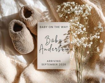 May include: A beige announcement card with the text "Baby on the Way" and "Baby Anderson, Arriving September 2026" is displayed next to a pair of knitted baby booties. A bouquet of white flowers rests on a beige knitted blanket.