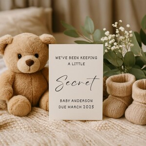 May include: A beige announcement card with the text "WE'VE BEEN KEEPING A LITTLE Secret BABY ANDERSON DUE MARCH 2025" is displayed next to a teddy bear and baby booties. The background includes greenery and a soft blanket.