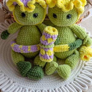 May include: Two handmade crochet dolls in the shape of flowers. The dolls are green with yellow and purple accents. They have black button eyes and are sitting on a white, textured surface.