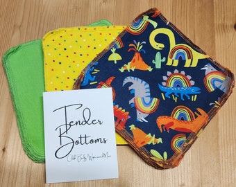Puede incluir: Un conjunto de tres toallitas para bebé de tela. Una toallita es amarilla con lunares rojos y blancos, una es verde y otra es azul con un patrón de dinosaurio y arcoíris. Las toallitas están dobladas y apiladas sobre una superficie de madera. El texto "Tender Bottoms Cloth Baby Wipes and More" está impreso en una tarjeta blanca.