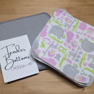 Puede incluir: Un montón de toallitas para bebés con un estampado de elefantes grises, jirafas rosas y cocodrilos verdes sobre un fondo blanco. También hay una toallita gris y una tarjeta con el texto "Tender Bottoms".