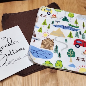 Puede incluir: Un pañal de tela blanco con un estampado colorido de escena de camping, un pañal de tela marrón y una tarjeta con el texto "Tender Bottoms". La escena de camping incluye una caravana roja, árboles y un lago.