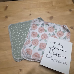 Puede incluir: Un montón de toallitas para bebés con un estampado floral de rosas y un estampado de lunares verdes. Una tarjeta blanca con las palabras "Tender Bottoms" está al lado de las toallitas.