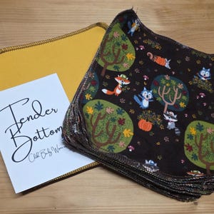 Puede incluir: Un montón de toallitas para bebés de color marrón oscuro con un estampado de animales del bosque, con zorros, mapaches y búhos. También hay un paño amarillo y una tarjeta con el texto "Tender Bottom".