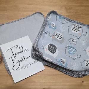 Puede incluir: Toallitas de tela para bebé con un estampado de elefantes en azul claro y bordes grises. Una pila de toallitas está junto a una toallita gris individual y una tarjeta blanca con el texto "Tender Bottom".