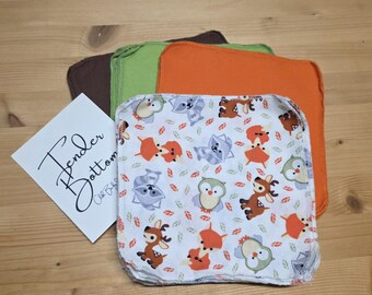 Puede incluir: Una pila de toallitas para bebés con un estampado de animales del bosque, con mapaches, ciervos y búhos sobre un fondo blanco. Toallitas de colores lisos en marrón, verde y naranja están apiladas detrás de las estampadas. Una tarjeta blanca con el texto "Tender Bottom" está a la izquierda.