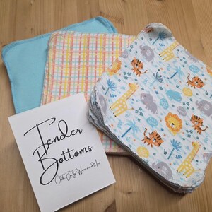 Puede incluir: Un conjunto de toallitas para bebés con un estampado de animales de safari. La imagen también muestra una toallita azul claro, una toallita a cuadros y una tarjeta con el texto "Tender Bottoms".
