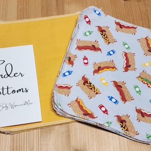 Puede incluir: Paños para bebés con un estampado de perritos calientes sobre un fondo azul claro. Junto a ellos, un paño amarillo y una tarjeta blanca con el texto "Tender Bottoms".