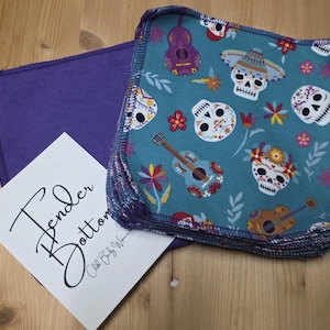 Puede incluir: Un conjunto de cuadrados de tela turquesa con un patrón de calaveras y guitarras del Día de Muertos, junto a un cuadrado morado liso y una tarjeta blanca con las palabras "Tender Bottoms". Los cuadrados de tela tienen un borde cosido.