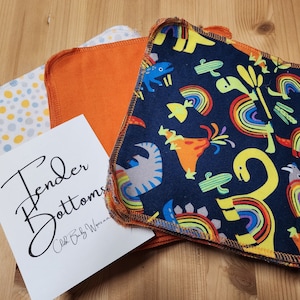 Puede incluir: Un montón de toallitas para bebé de tela con una variedad de patrones coloridos, incluyendo un estampado de dinosaurio azul y amarillo, un estampado de lunares amarillo y blanco y un estampado naranja sólido. Las toallitas están etiquetadas como "Tender Bottoms Cloth Baby Wipes".