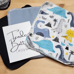 May include: A stack of black, blue and white reusable cloth baby wipes with a dinosaur print. The wipes are folded and stacked on a wooden surface. The text "Tender Bottoms Cloth Baby Wipes" is printed on a white card.