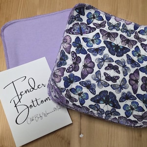 Puede incluir: Un conjunto de toallitas para bebé. Una es lisa, de color lavanda, y la otra está adornada con un estampado de mariposas azules y moradas. También hay una tarjeta blanca con la inscripción "Tender Bottom".
