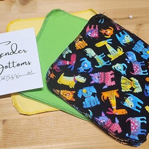 Puede incluir: Un conjunto de tres paños reutilizables, uno negro con un patrón de perros coloridos, uno verde y uno amarillo. Los paños están doblados y apilados sobre una superficie de madera. El texto "Tender Bottoms" está escrito en una tarjeta blanca delante de los paños.