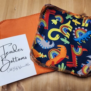May include: A stack of reusable cloth baby wipes with a dinosaur and rainbow print on a dark blue background. The wipes are surrounded by a light orange fabric. The text "Tender Bottoms Cloth Baby Wipes and More" is printed on a white card.