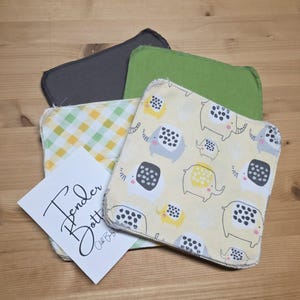 May include: A selection of baby washcloths in different colours and patterns. One cloth has a yellow background with elephant illustrations. Other cloths are solid grey, green, and a yellow and green gingham pattern. A white card with the text "Tender Bott" is also included.
