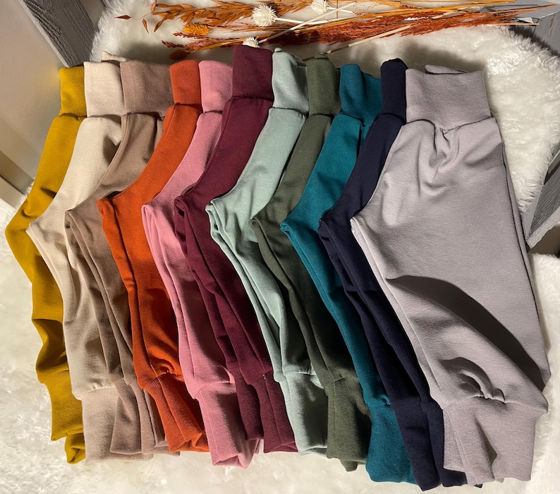 Puede incluir: Una colecci&oacute;n de pantalones coloridos con cinturas altas y dobladas y tobillos con pu&ntilde;os. Los pantalones son de varios tonos, incluyendo amarillo mostaza, beige, naranja, rosa, burdeos, verde, verde azulado, azul marino y gris. Los pantalones est&aacute;n dispuestos en fila.