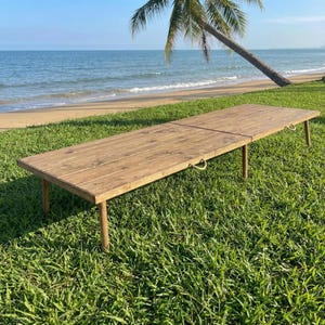 May include: A long, rectangular wooden table with a natural finish, set on four legs. The table is outdoors on a grassy area, with a beach and ocean in the background. Rope handles are on the sides.