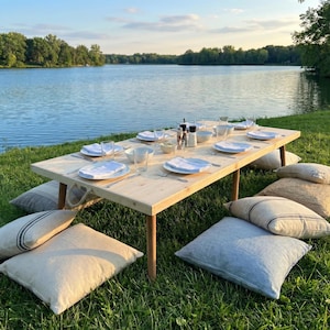 May include: A wooden picnic table set for a meal, with plates, glasses, and silverware. The table is surrounded by pillows on the grass. The scene is set near a lake with trees in the background, creating a serene outdoor dining experience.