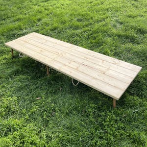 Puede incluir: Una mesa de madera larga y rectangular con un acabado natural, sostenida por cuatro patas delgadas. La mesa tiene asas de cuerda en cada extremo y est&aacute; sobre hierba verde. La mesa mide aproximadamente 183 cm de largo.