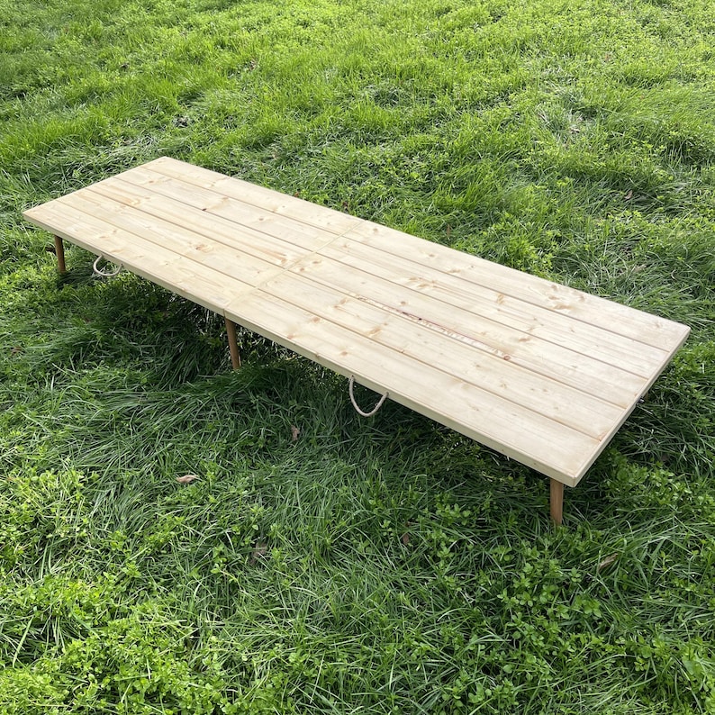 Puede incluir: Una mesa de madera larga y rectangular con un acabado natural, sostenida por cuatro patas delgadas. La mesa tiene asas de cuerda en cada extremo y est&aacute; sobre hierba verde. La mesa mide aproximadamente 183 cm de largo.