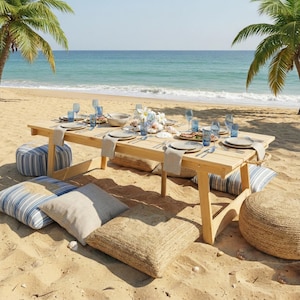 May include: A beach picnic setup with a light wood picnic table set with plates, glasses, and silverware. Blue and white striped cushions and beige pillows surround the table. The ocean and palm trees are in the background.