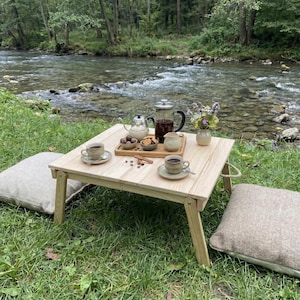 May include: A light wood folding table set for a picnic, with coffee cups, a teapot, and snacks. Two beige pillows are on the grass. A river flows in the background, surrounded by trees.