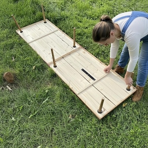 Puede incluir: Una mesa de madera de color claro con cuatro patas se est&aacute; montando al aire libre sobre una superficie cubierta de hierba. La mesa tiene forma rectangular con una tapa de listones y ribete marr&oacute;n oscuro. Las patas son cil&iacute;ndricas.