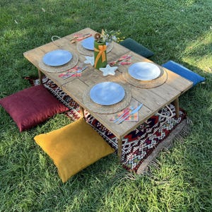 May include: A low wooden picnic table set for a meal on the grass. The table has four blue plates, woven placemats, and colorful napkins. Pillows in various colors surround the table, and a decorative rug is underneath.