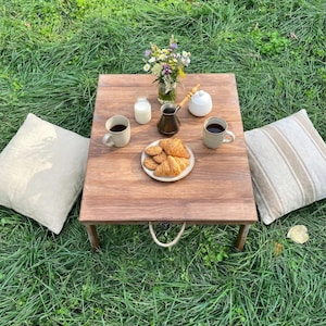 May include: A wooden picnic table set for a relaxing outdoor meal. The table holds coffee, croissants, cookies, milk, and a vase of flowers. Two beige pillows are on the grass, creating a cozy setting.