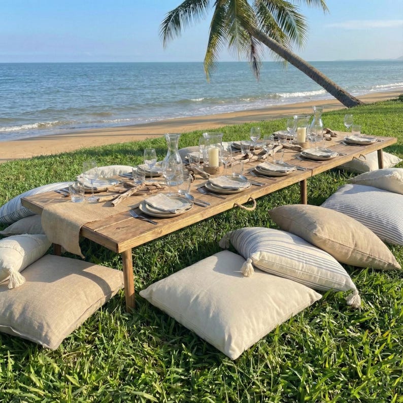 Puede incluir: Una mesa r&uacute;stica de madera preparada para un picnic en la playa, con platos, vasos y velas. Cojines beige est&aacute;n esparcidos alrededor de la mesa sobre la hierba verde. El oc&eacute;ano y una palmera est&aacute;n en el fondo.