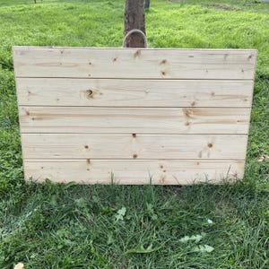 Puede incluir: Un panel rectangular de madera, construido con tablones horizontales de pino sin terminar. La veta de la madera y los nudos son visibles. El panel se encuentra sobre un fondo de hierba verde y un tronco de &aacute;rbol.