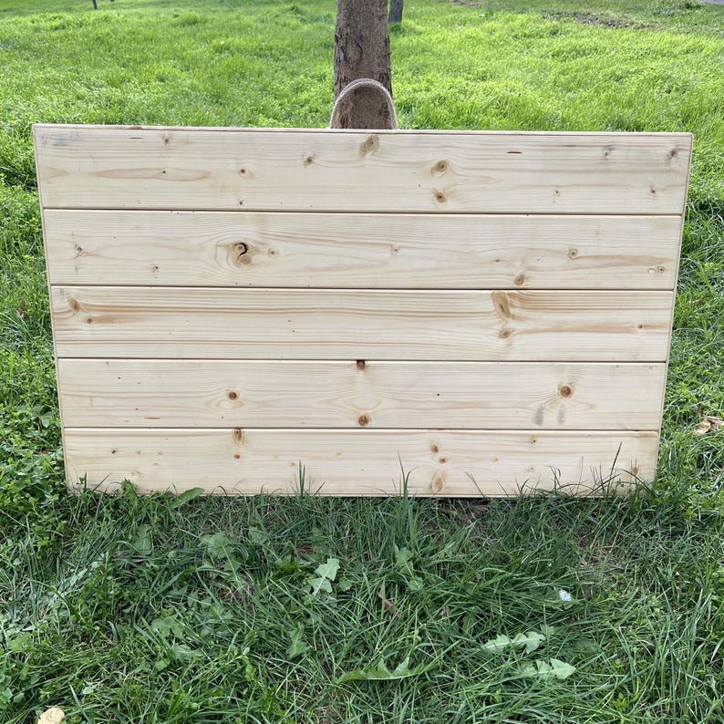 Puede incluir: Un panel rectangular de madera, construido con tablones horizontales de pino sin terminar. La veta de la madera y los nudos son visibles. El panel se encuentra sobre un fondo de hierba verde y un tronco de &aacute;rbol.