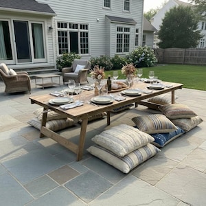 May include: A rustic wooden outdoor dining table set for a meal, surrounded by floor cushions. The table is set with plates, glasses, and a breadboard. The scene is set on a stone patio with a house and yard in the background.