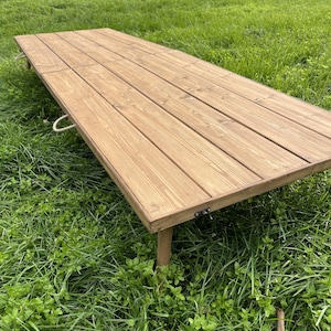 Könnte beinhalten: Ein rechteckiger, klappbarer Holztisch mit hellbraunem Finish. Der Tisch besteht aus Holzbrettern und hat Metallscharniere. Er hat zwei Seilgriffe und steht auf einer grasbewachsenen Oberfläche.