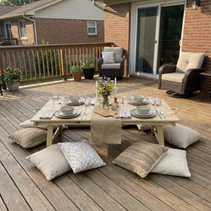 May include: A low, light-coloured wooden picnic table is set for a meal on a wooden deck. The table is surrounded by cushions and set with plates, bowls, cutlery, and glasses. A vase of flowers and candles complete the setting.