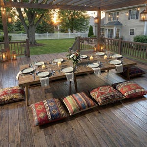 May include: Outdoor dining setting with a rustic wooden table set for a meal. The table is surrounded by floor cushions with colorful patterns. Overhead, a wooden pergola is adorned with string lights, creating a warm ambiance.