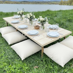 May include: A picnic setting with a light wood table, white plates, and glassware. Cream-colored cushions are arranged on the grass. White flowers in vases and candles decorate the table. The scene is set near a lake.