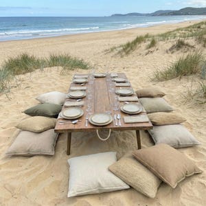 Peut inclure: Une table en bois rustique dressée pour un pique-nique sur la plage, entourée de coussins beiges et vert olive. La table est garnie d'assiettes, de couverts et de verres. La plage et l'océan sont en arrière-plan.