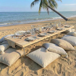 May include: A beach picnic setup with a long wooden table set with plates, glasses, and candles. Beige pillows surround the table on the sand. A palm tree and the ocean are in the background.