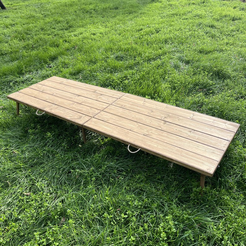 Puede incluir: Una mesa plegable rectangular de madera con acabado marr&oacute;n claro. La mesa est&aacute; hecha de tablones de madera y tiene patas de metal. La mesa est&aacute; colocada sobre una superficie cubierta de hierba. La mesa mide aproximadamente 183 cm de largo.