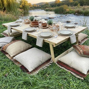 Peut inclure: Une table de pique-nique en bois dressée pour un repas en plein air, avec des assiettes, des verres et des couverts. Des coussins et des couvertures sont posés sur le sol. La table est installée près d'une rivière.