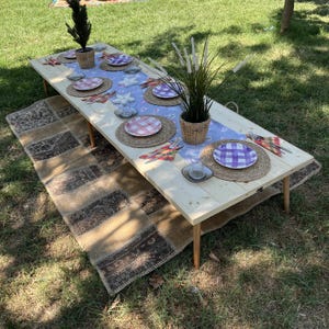 Puede incluir: Una larga mesa de picnic de madera clara preparada para una comida al aire libre. La mesa est&aacute; puesta con platos, manteles individuales y cubiertos. Una alfombra estampada est&aacute; debajo de la mesa. Plantas decorativas en cestas tejidas y un camino de mesa azul completan el escenario.
