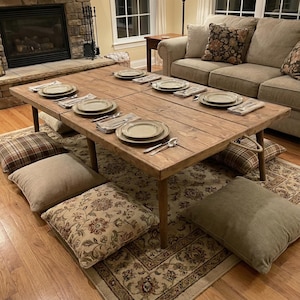 May include: A large, rectangular wooden table set for a meal, with place settings for eight people. The table is surrounded by floor cushions and a patterned rug. The table is approximately 72 inches long.