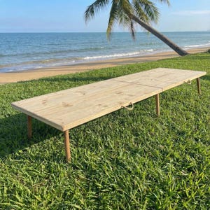 May include: A long, rectangular wooden table with four legs and rope handles. The table is made of light-colored wood and is set on a grassy area near a beach with a palm tree and ocean in the background.