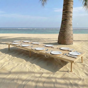 May include: A long, light-colored wooden table set for a meal on a sandy beach. White plates, silverware, and napkins are arranged for multiple place settings. A palm tree trunk is visible in the background, with the ocean and sky in the distance.
