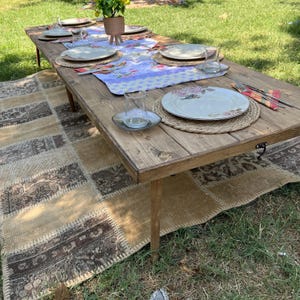 Puede incluir: Una mesa de picnic r&uacute;stica de madera preparada para una comida al aire libre. La mesa est&aacute; rodeada por una alfombra estampada y hierba verde. La mesa est&aacute; puesta con platos, vasos y un centro de mesa floral.